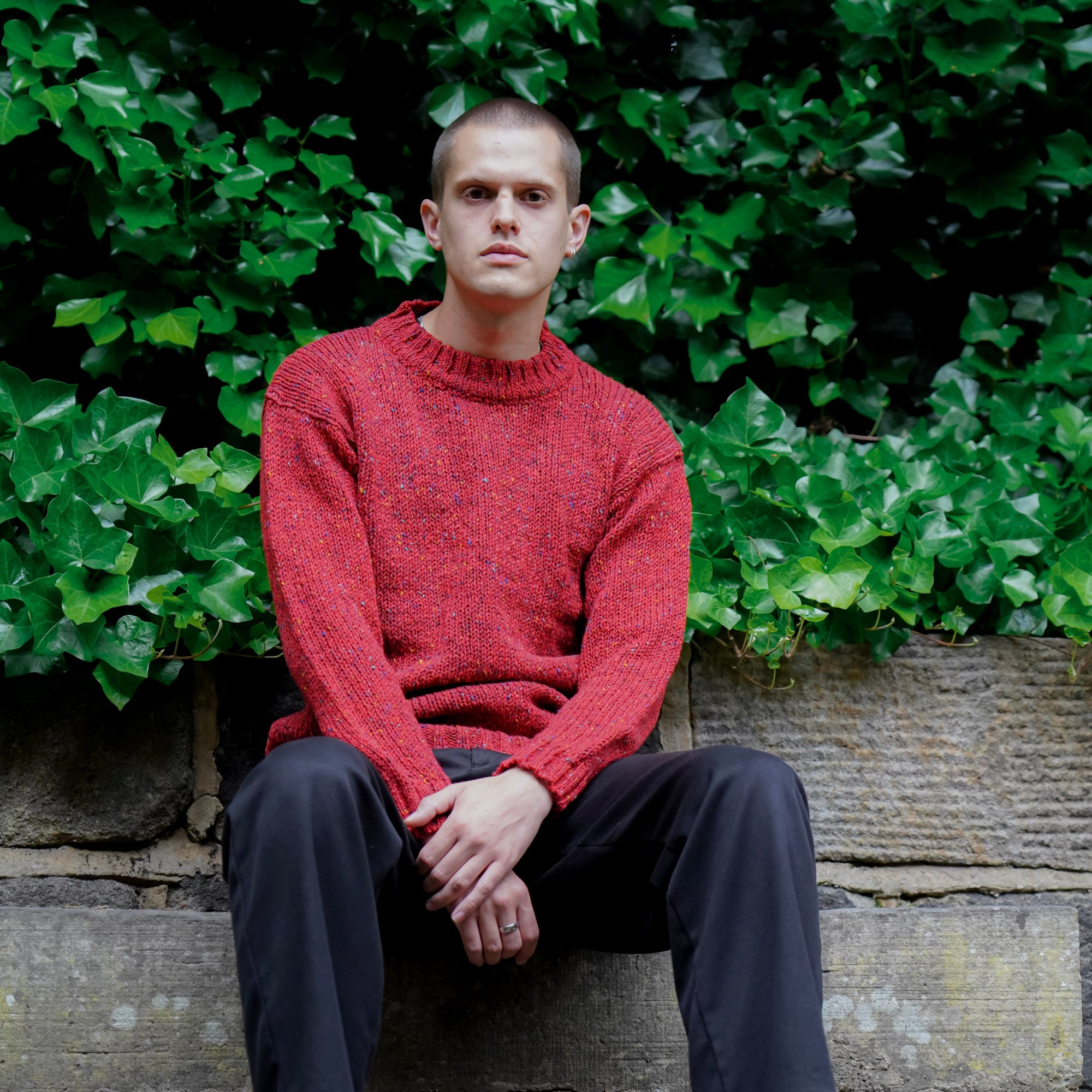 A man sits on stone steps outdoors in front of dense green ivy, wearing a textured red tweed wool knit sweater, dark trousers, and black shoes.