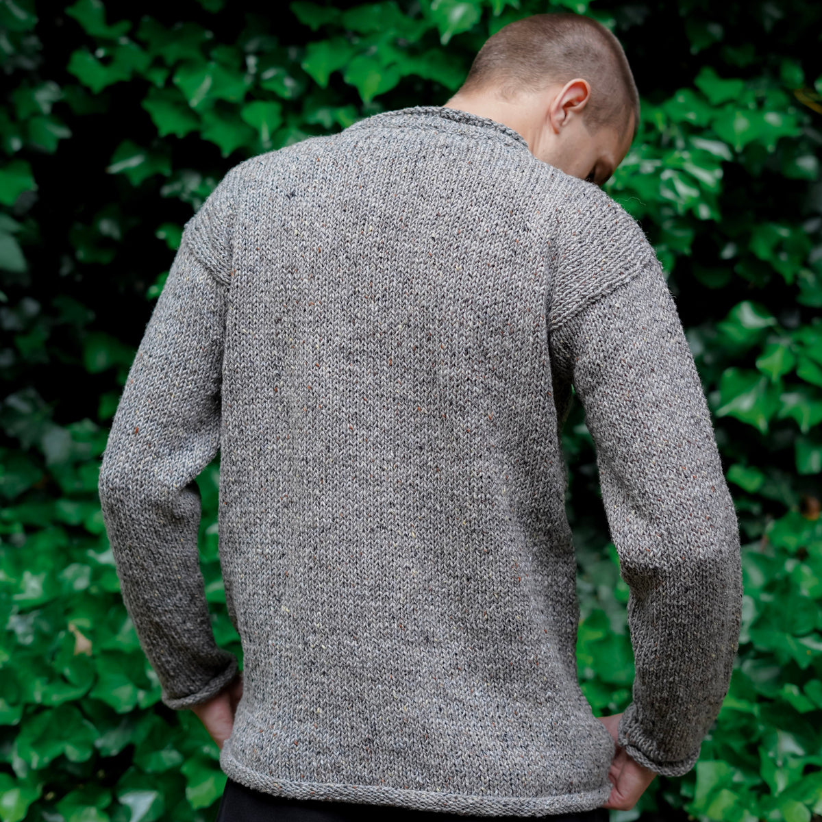 A man viewed from behind wearing a textured gray knit sweater and black trousers, standing outdoors against a background of green ivy.