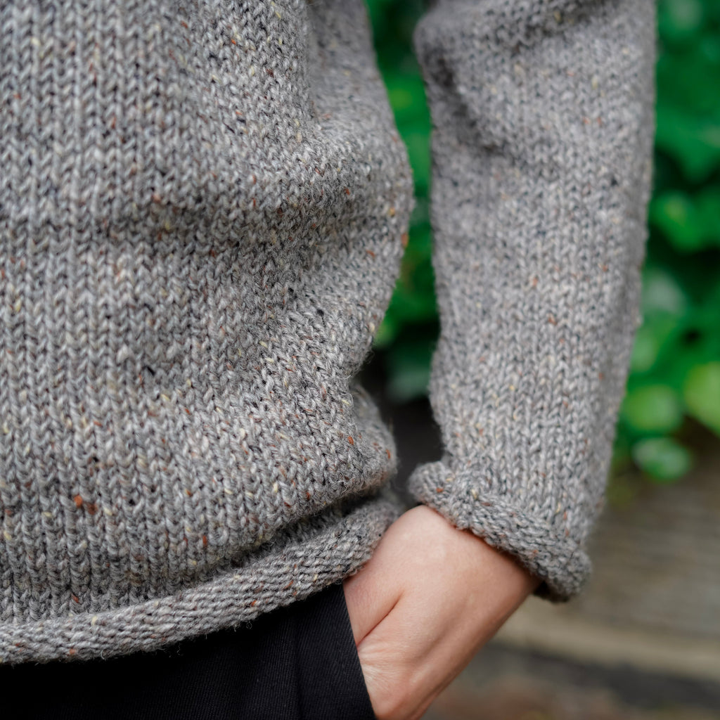 Close-up of a person wearing a chunky gray knit sweater with a hand in the pocket of black trousers, with a blurred green outdoor background.