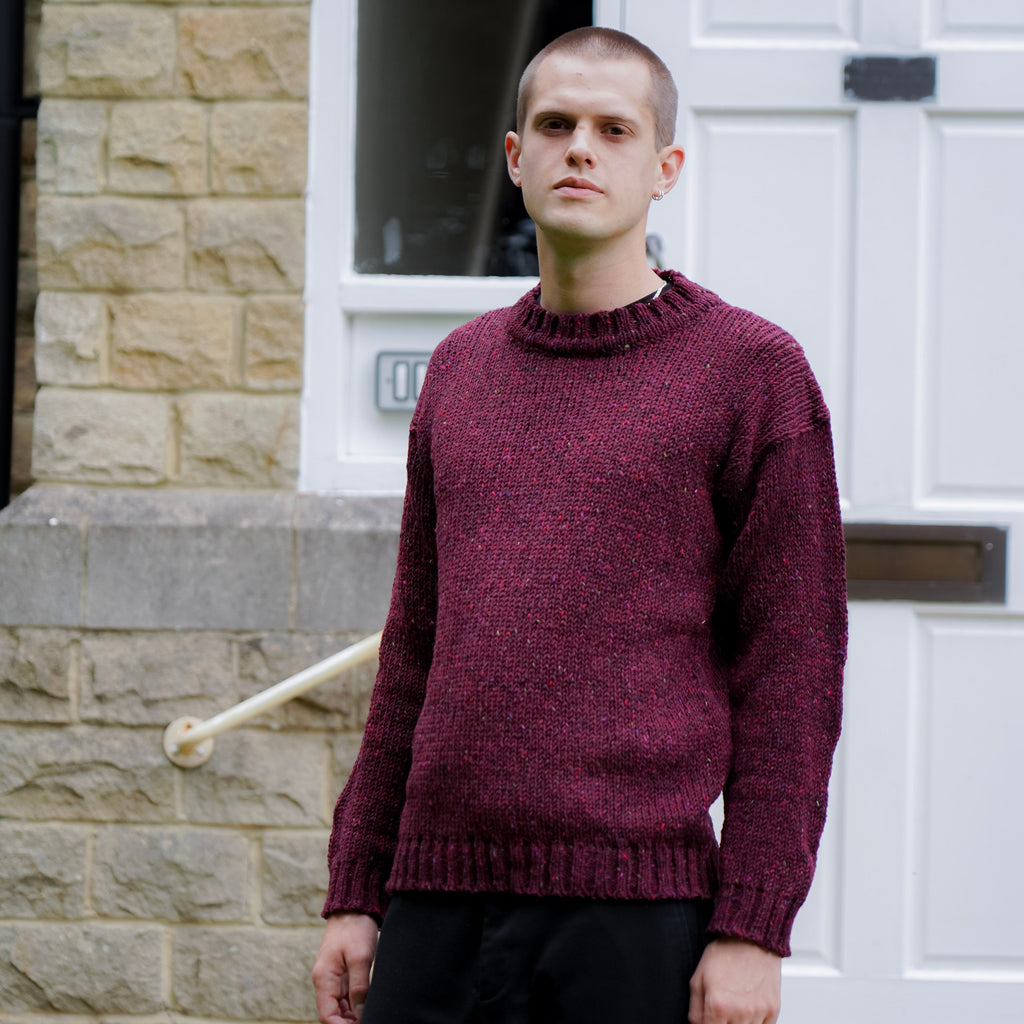 A man wearing a maroon knit crewneck sweater and black trousers, standing in front of a white door and stone wall.