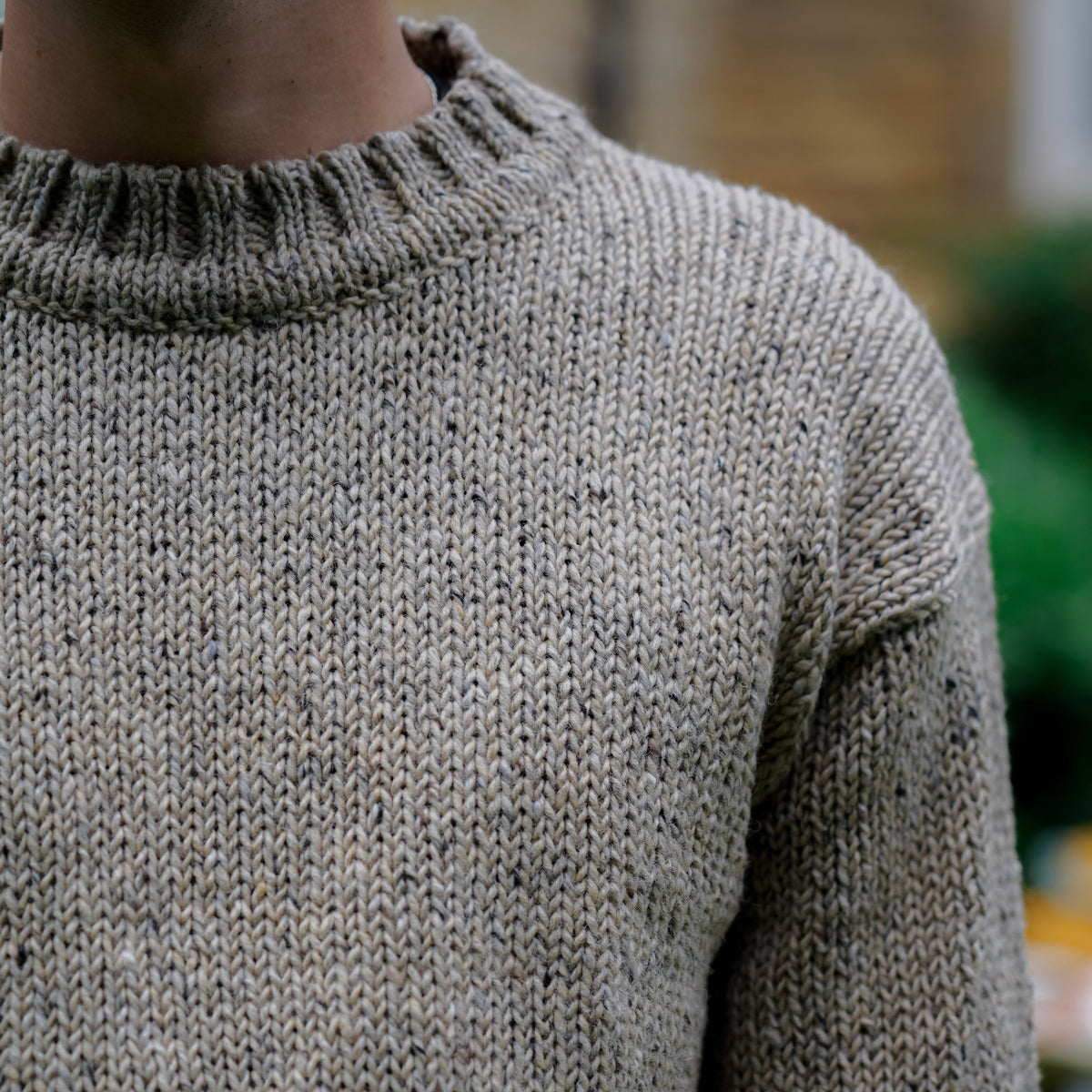 Close-up of a person wearing a chunky knit crewneck sweater in a heathered taupe-gray color, showing the ribbed collar and textured knit fabric.