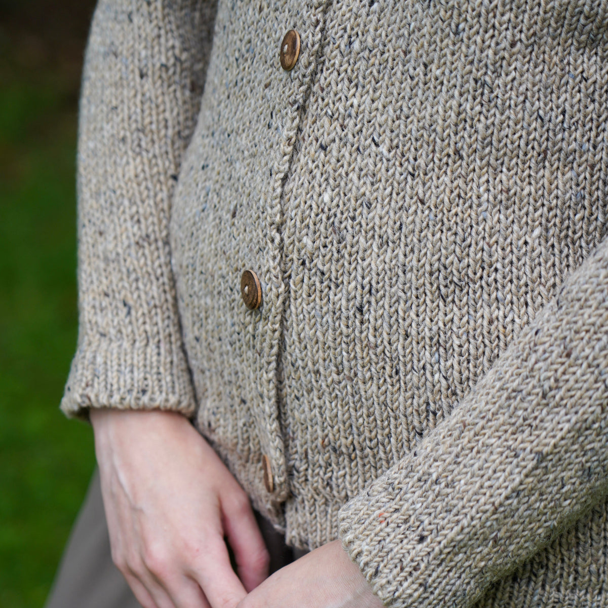 Close-up of a person wearing a chunky knit cardigan in a heathered oatmeal-beige color with brown buttons, paired with taupe trousers, set against a soft blurred green outdoor background.