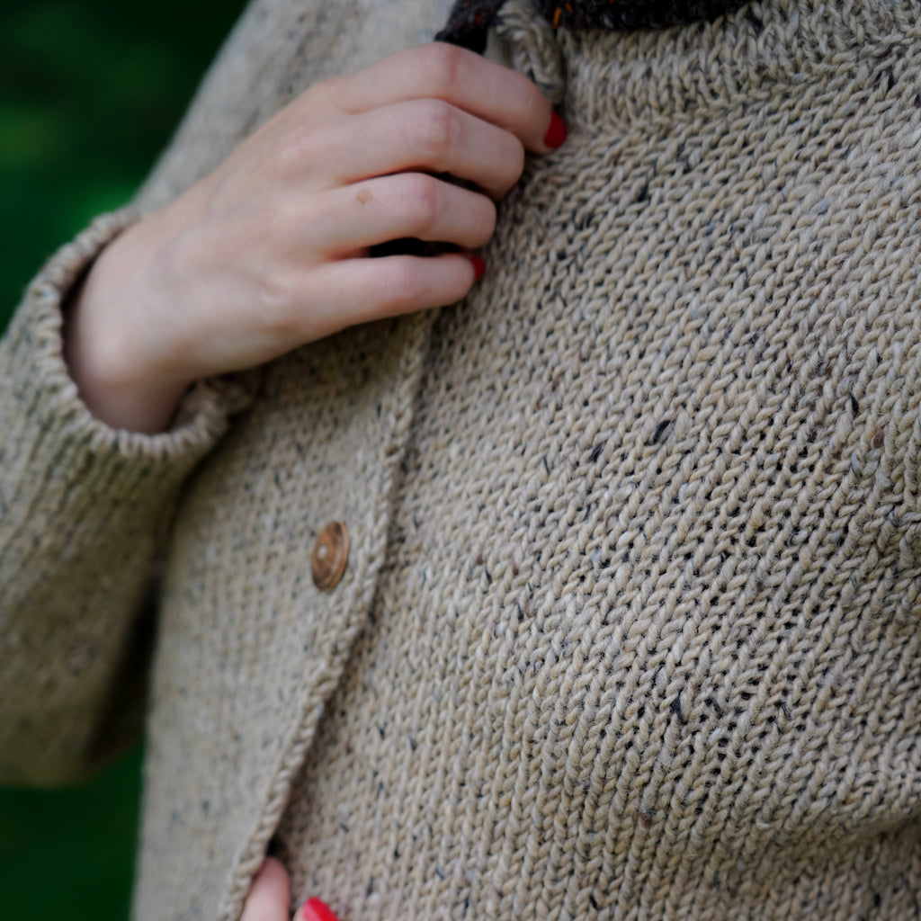 Close-up of a person wearing a chunky knit cardigan in a warm taupe-beige color with small dark flecks and brown buttons, with hands adjusting the collar and holding the front open.