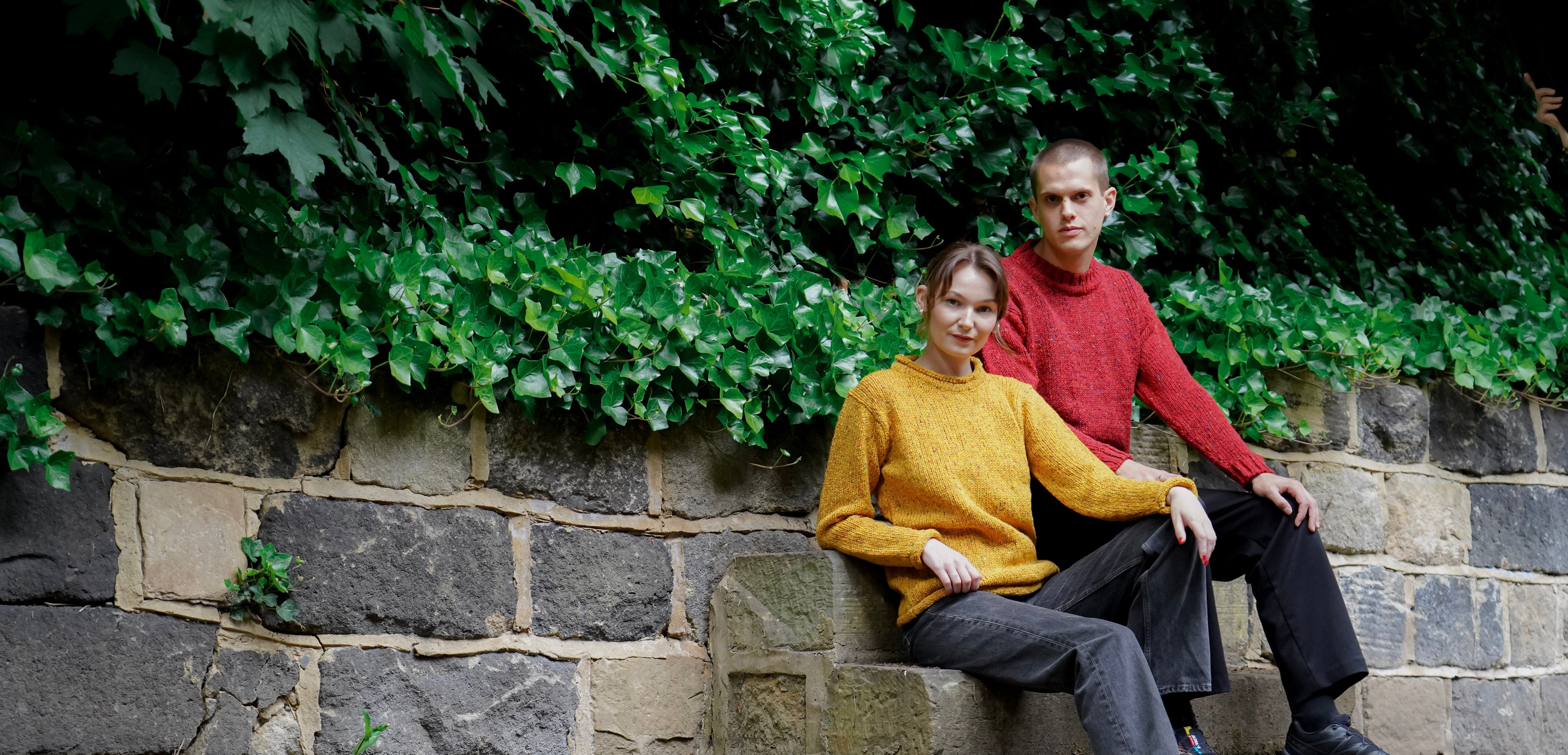 A man and a woman sitting together on a stone wall outdoors, with green ivy growing along the wall behind them. The woman wears a mustard yellow sweater, dark gray pants, and brown shoes, while the man wears a red sweater, dark pants, and black shoes.