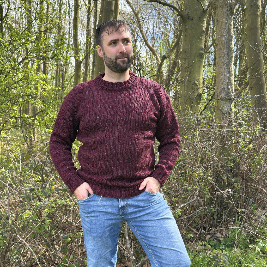 Man wearing a maroon sweater and blue jeans standing in a forest.
