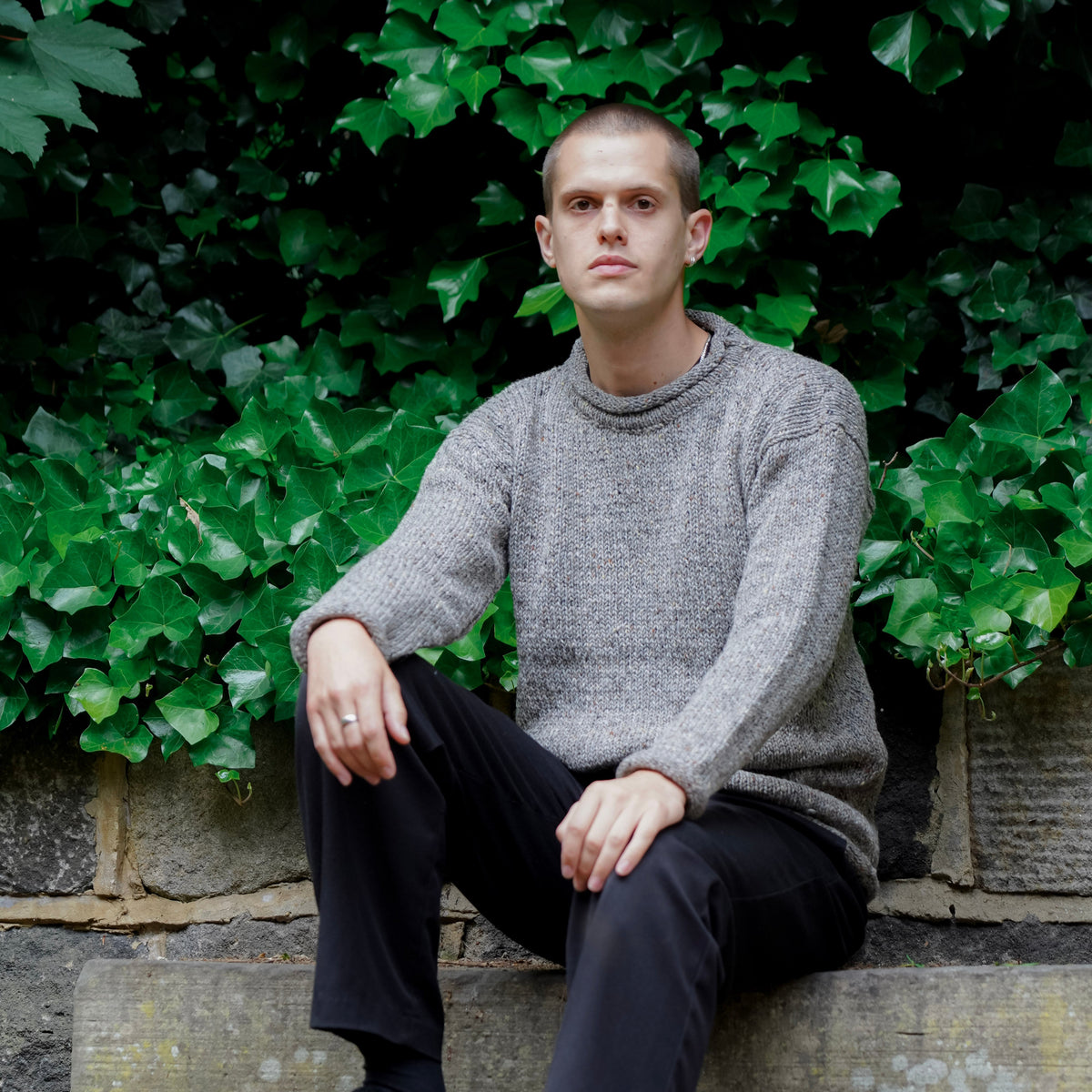 A man sits on stone steps outdoors, wearing a gray knit sweater, black trousers, and black shoes, with green ivy and a stone wall behind him.