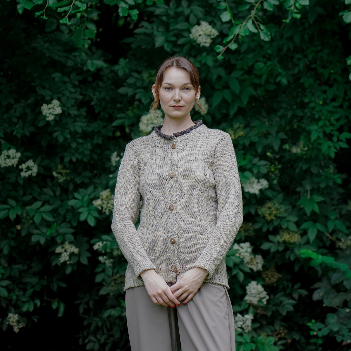 A woman stands outdoors in front of dense green foliage, wearing a textured light beige cardigan with dark trim and buttons, paired with matching beige trousers.