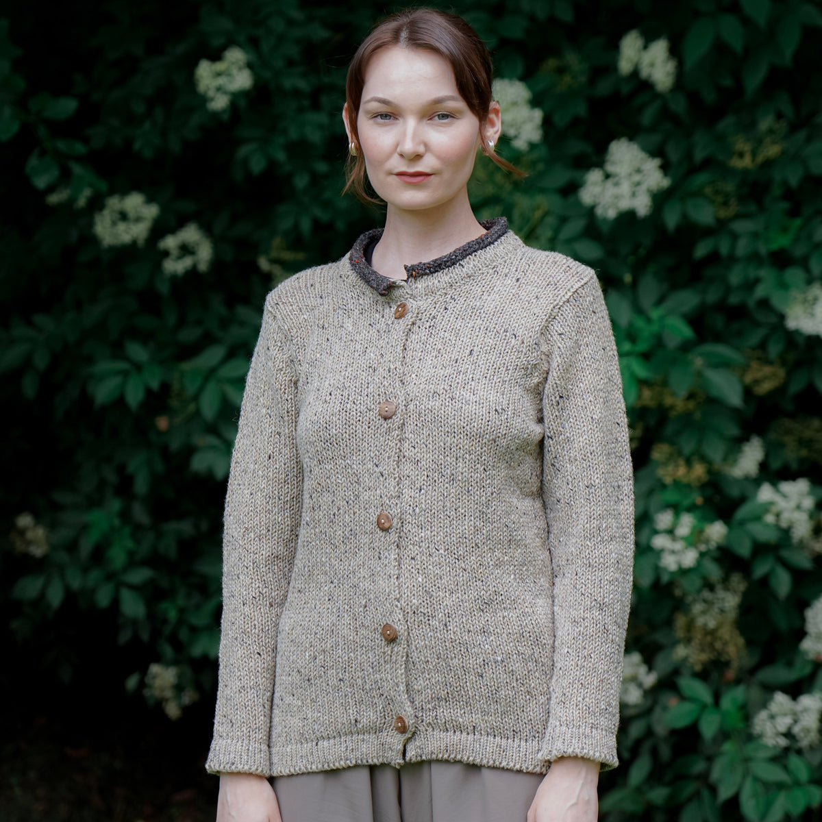 A woman stands outdoors in front of a dark green leafy wall with small white flowers, wearing a textured knit cardigan in a warm beige-gray color with brown buttons and a dark patterned collar, paired with taupe trousers.