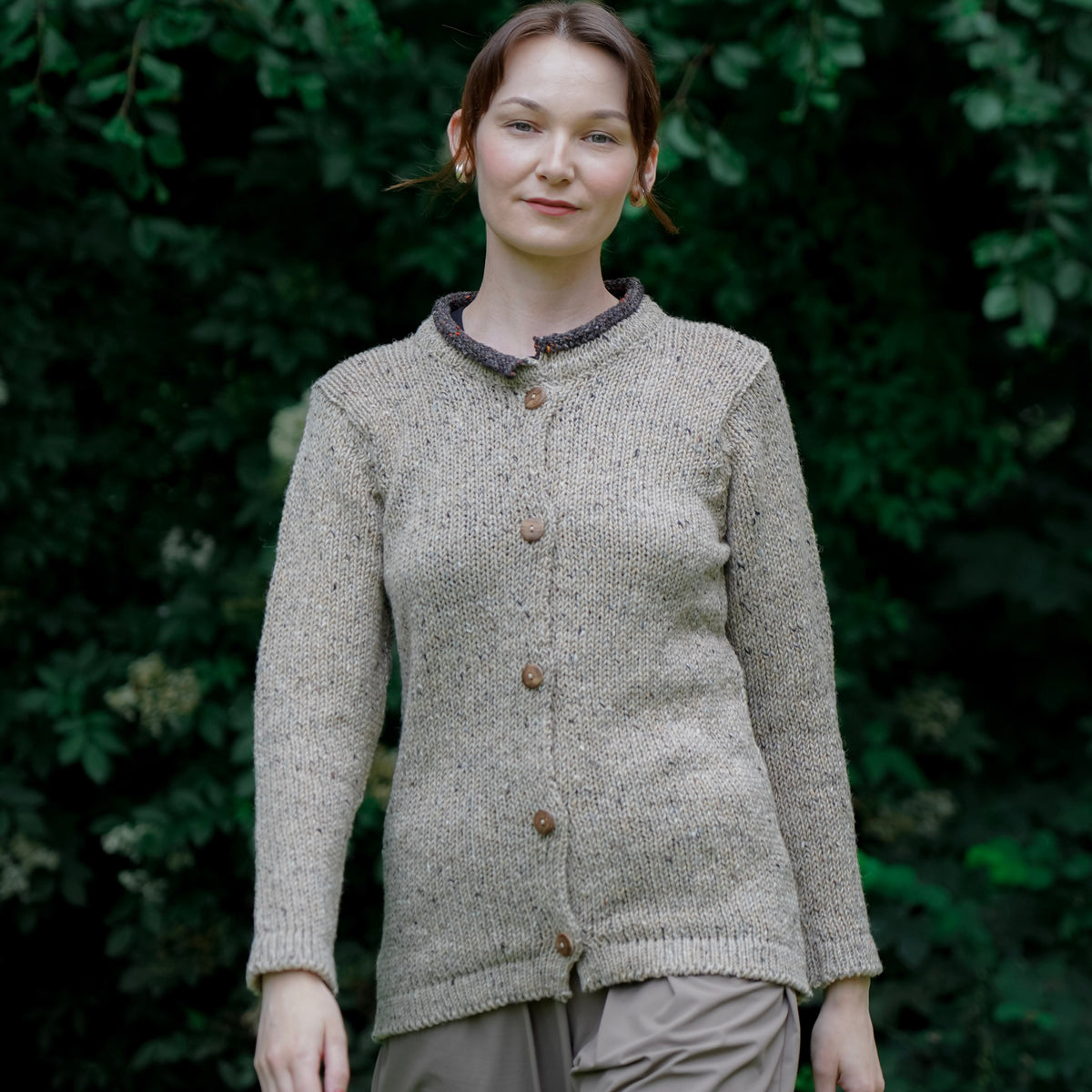 A woman wearing a light beige textured knit cardigan with brown buttons and a dark patterned collar, paired with olive green pants, standing against a background of dark green foliage.