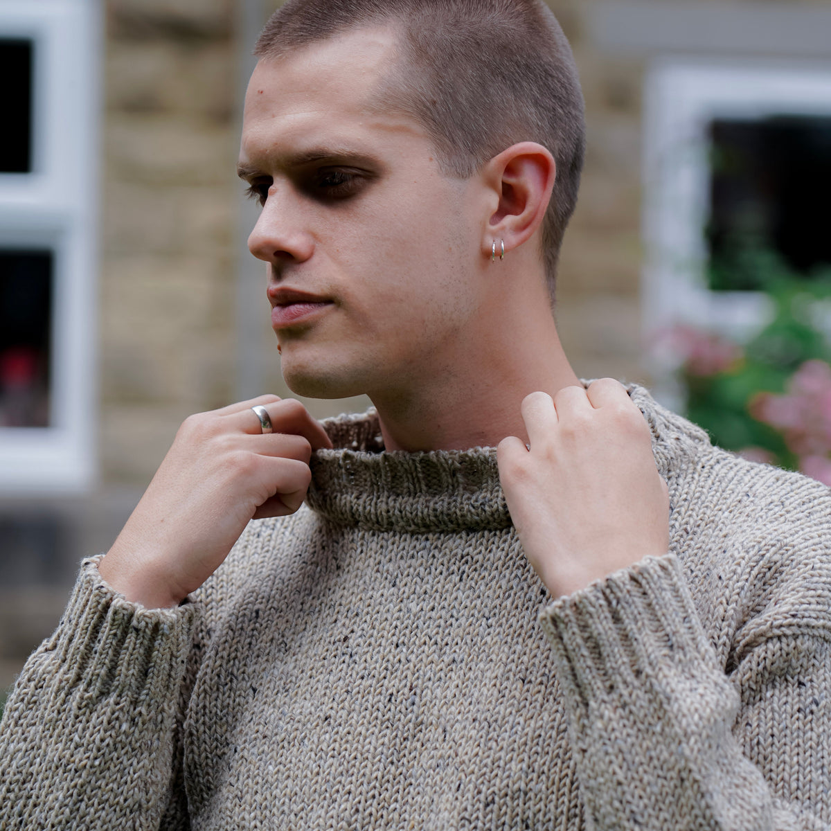 A man wearing a textured, speckled oatmeal-beige knit sweater, adjusting the collar with both hands. The background shows a softly blurred stone wall and windows.