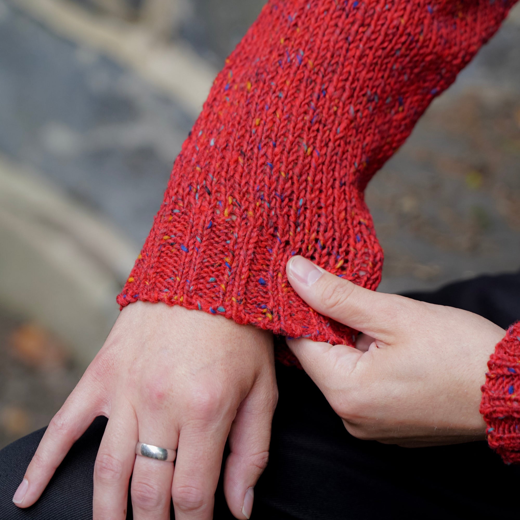 A close-up of a person wearing a textured red tweed wool knit crew sweater, showing the sleeve and hand, with another hand holding the cuff. The person is also wearing a silver band ring on their finger.