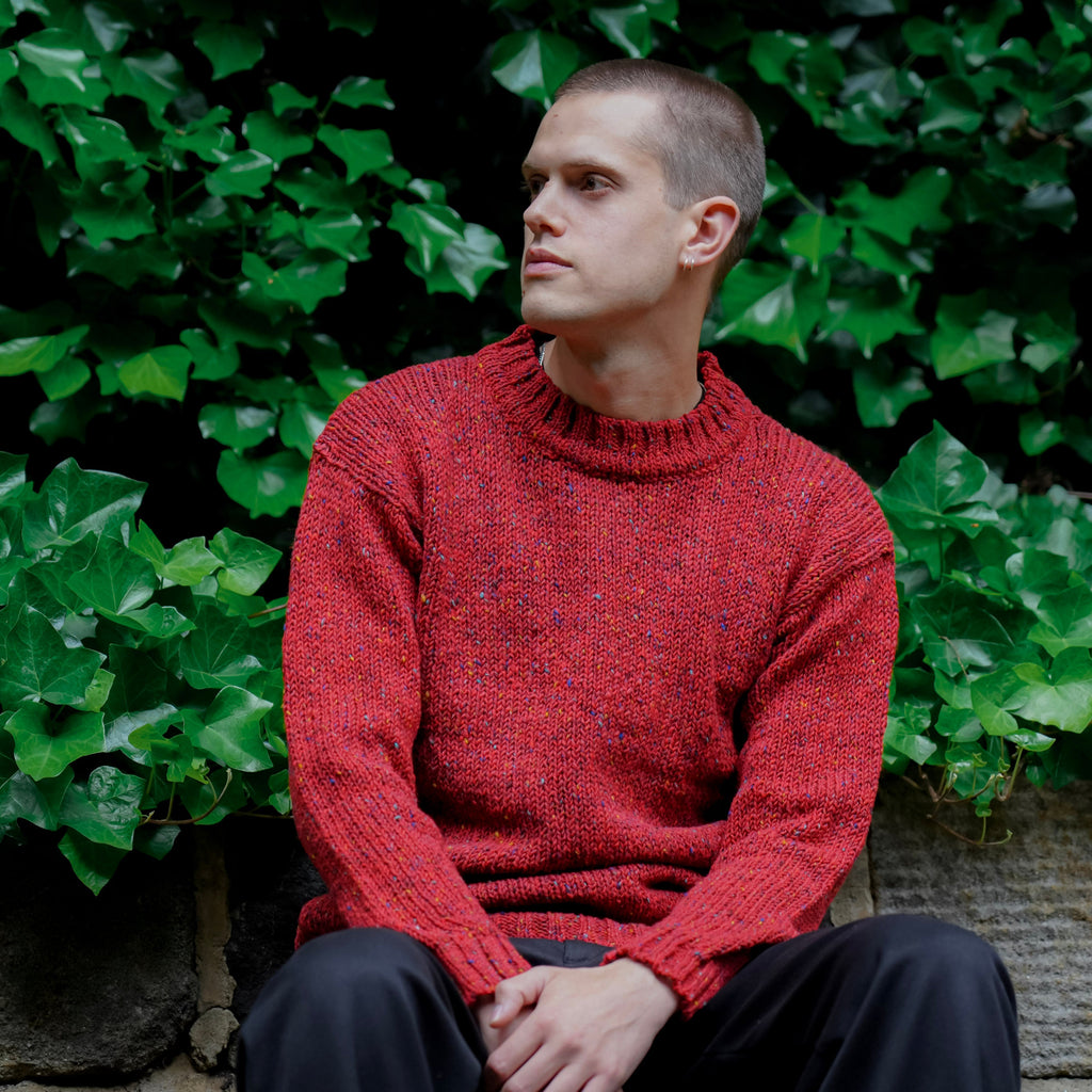 A man wearing a textured red knit mock-neck sweater and dark trousers sits on a stone wall in front of a dense background of green ivy.