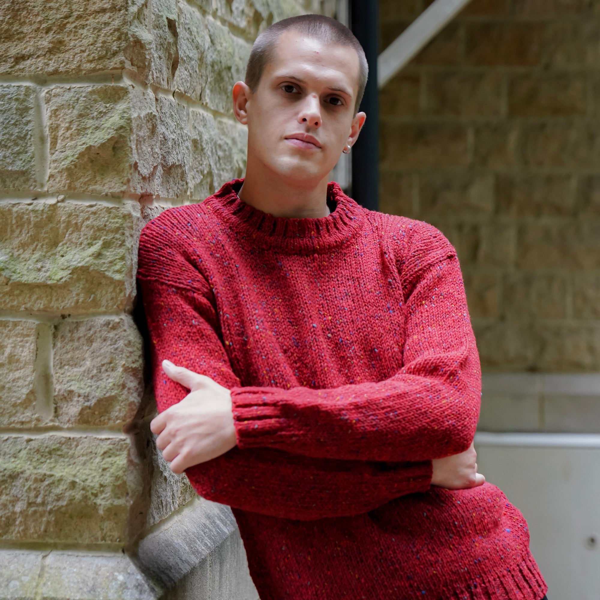 A man wearing a textured red knit crew neck sweater and black trousers, leaning against a stone brick wall.