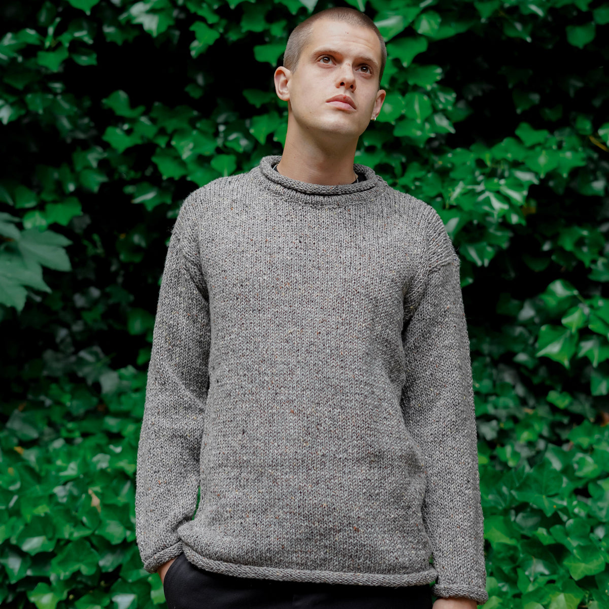 A man wearing a textured gray knit sweater and black pants, standing outdoors in front of a dense green ivy wall.