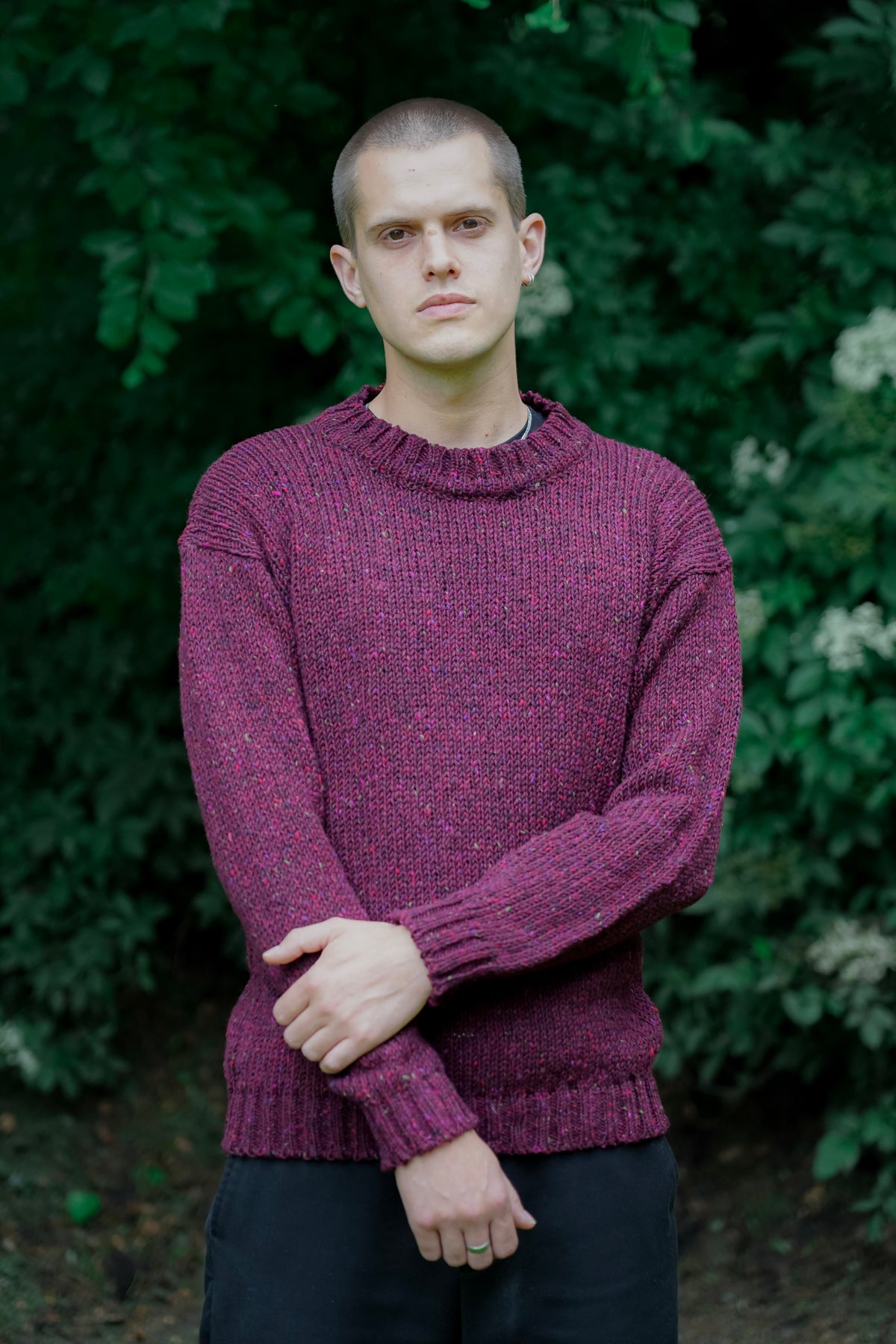 A man wearing a textured, heathered purple knit sweater and dark trousers, standing outdoors with his arms crossed in front of a background of green foliage.
