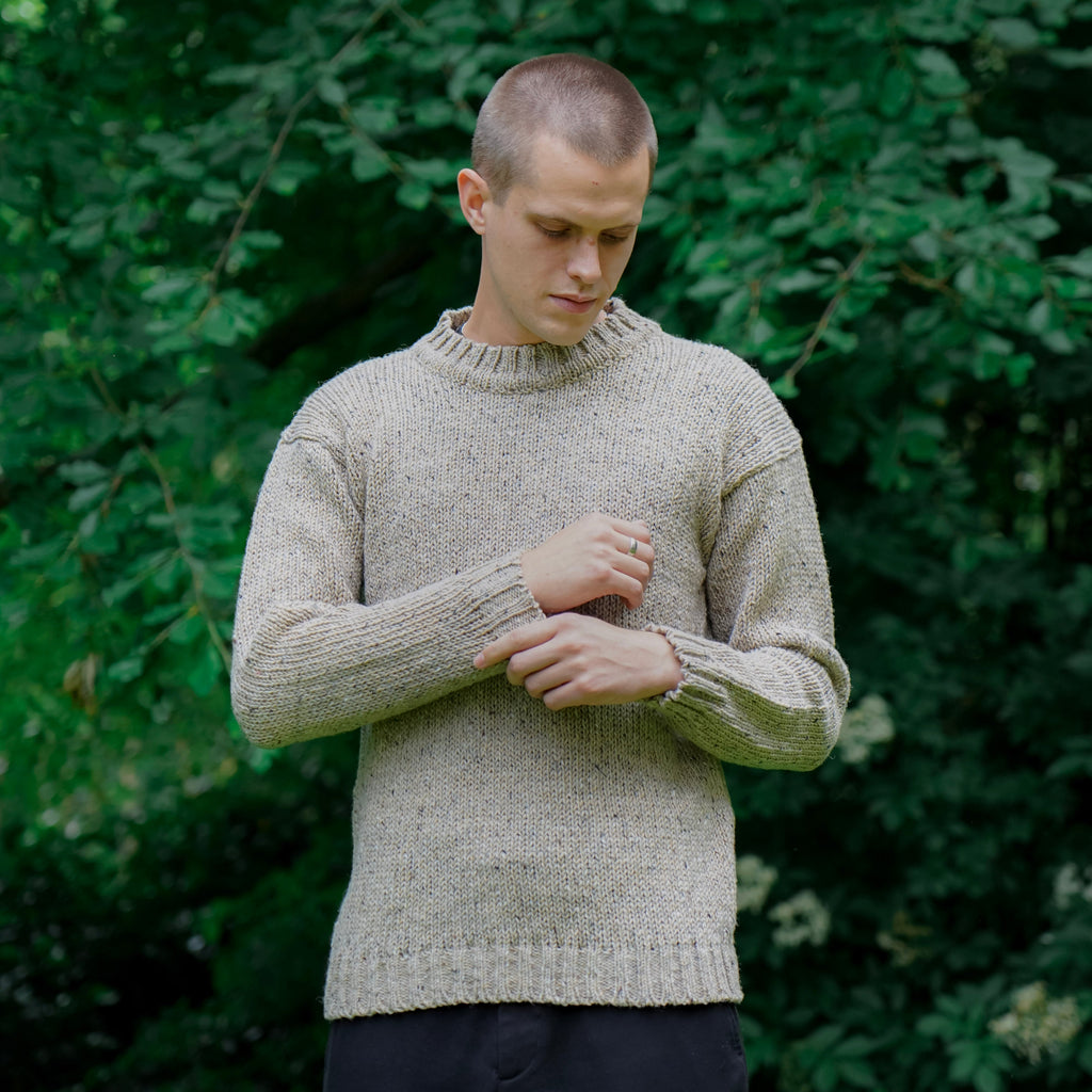 A man wearing a textured beige knit sweater and dark trousers standing outdoors in front of a dense green forest background.
