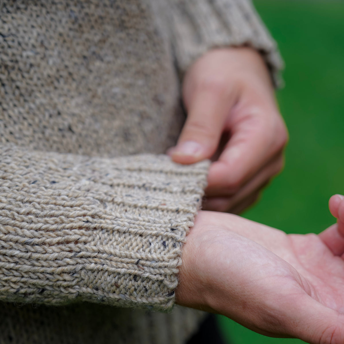 Close-up of a person wearing a chunky knit sweater, pulling the sleeve slightly to show the ribbed cuff texture against a blurred green outdoor background.