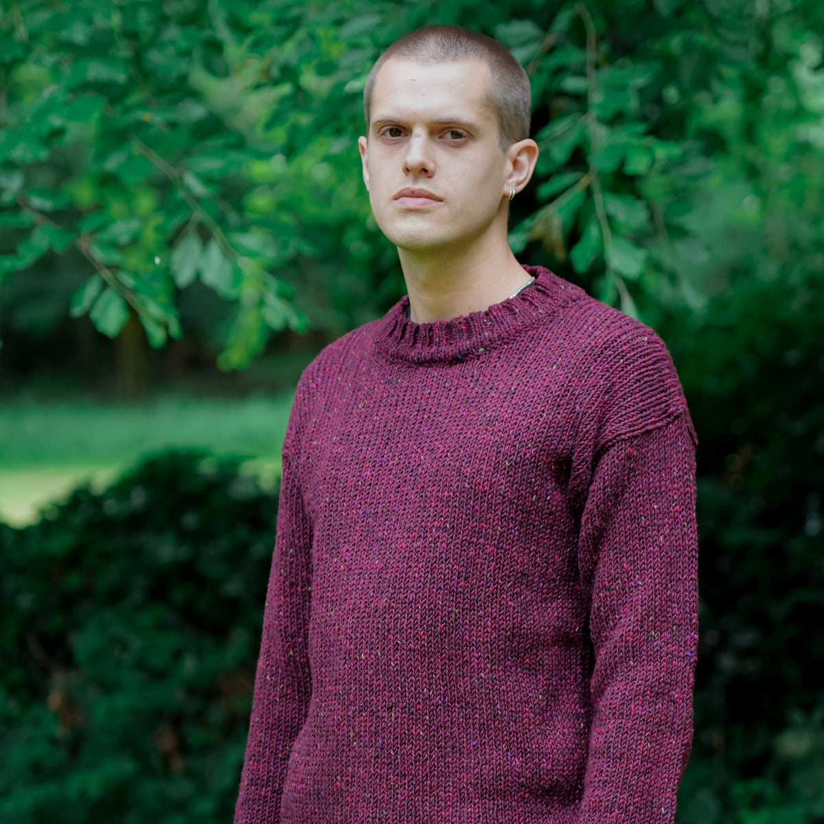 A man wearing a textured, heathered burgundy knit sweater and dark trousers, standing outdoors in front of a soft-focus background of green trees and bushes.