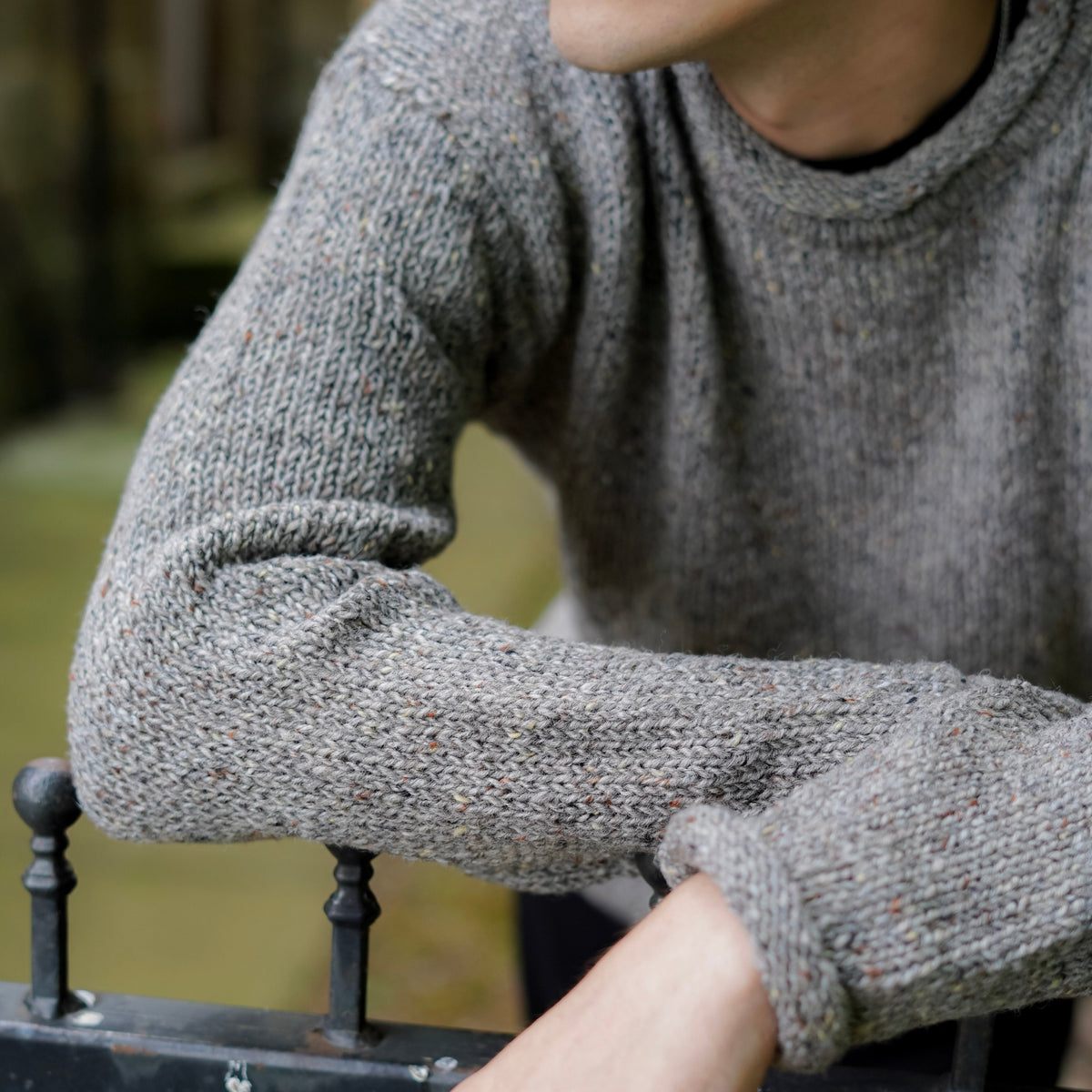 A close-up of a person wearing a textured gray knit crewneck sweater, leaning on a black metal railing in an outdoor setting.