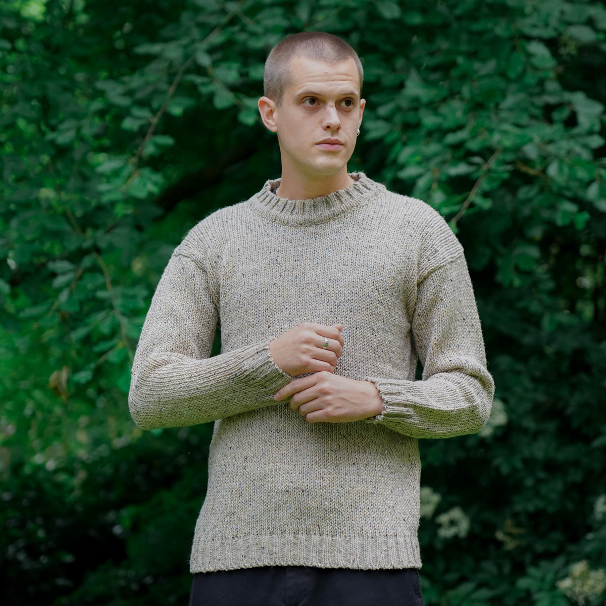 A man wearing a beige knit crewneck sweater and dark trousers, standing outdoors against a background of green trees.