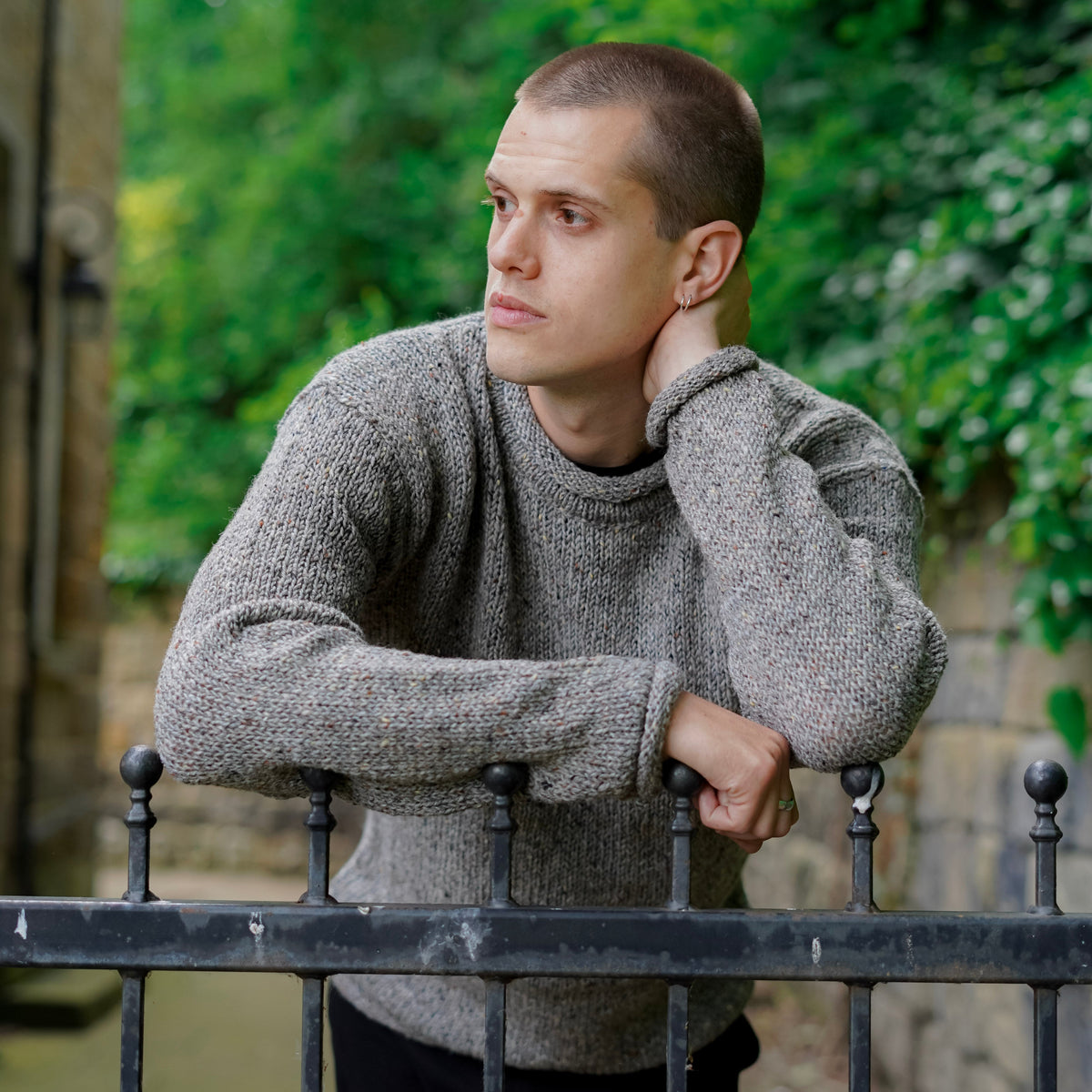 A man wearing a textured gray knit sweater and black trousers leans on a black metal fence in an outdoor garden setting with green foliage in the background.